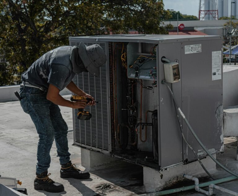 A worker in a bucket hat repairs an outdoor air conditioning unit on a rooftop.